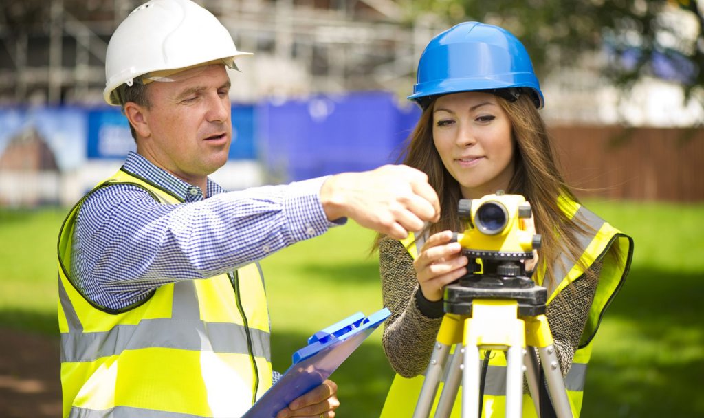 A man and a woman wearing hardhats and using a piece of surveying equipment.